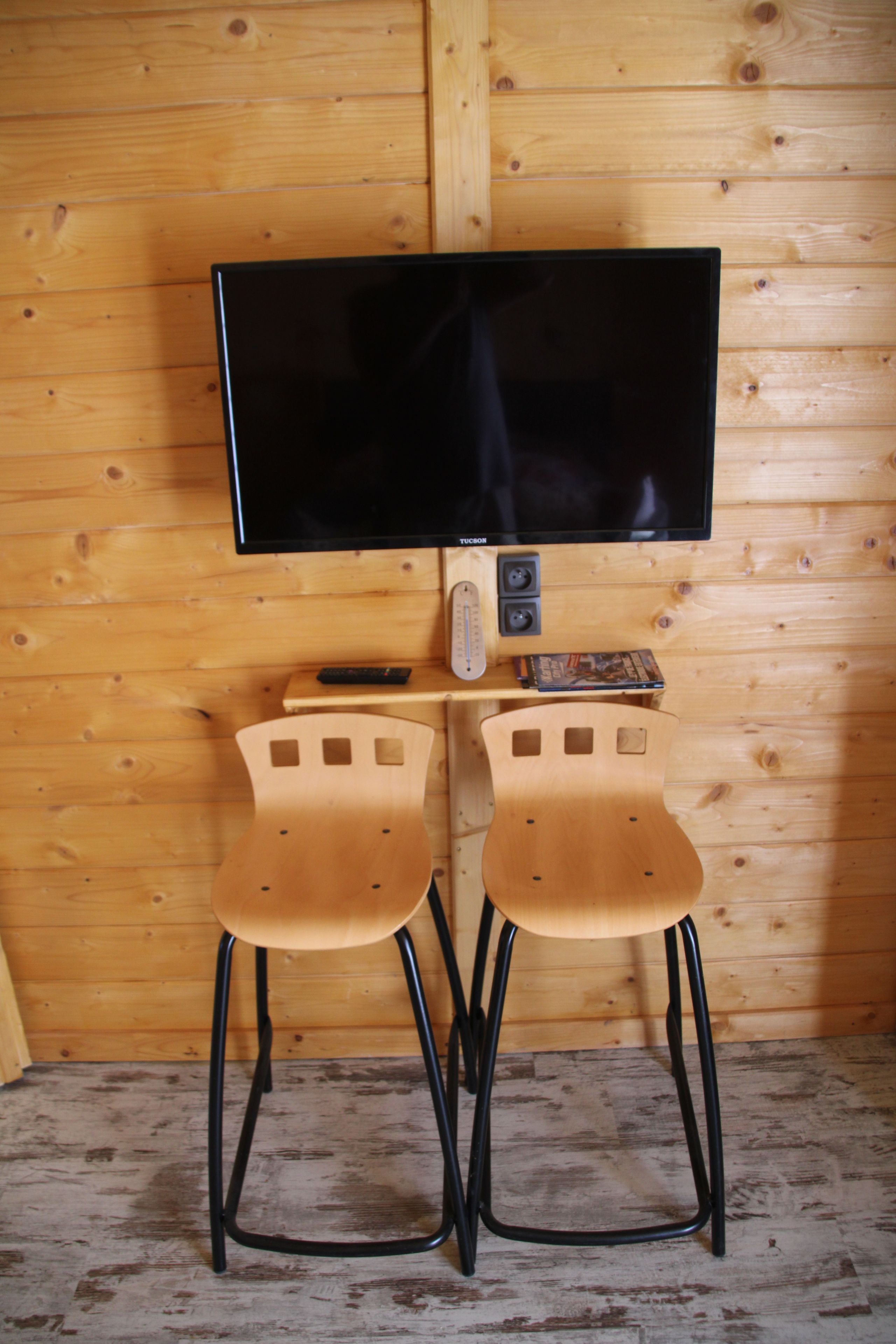 Een kamer met houten lambrisering, een flatscreen televisie bevestigd aan een houten voet, twee houten stoelen met zwarte poten en een klein tafeltje met een afstandsbediening en een boek.