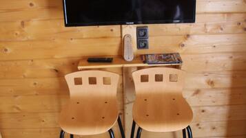 Een kamer met houten lambrisering, een flatscreen televisie bevestigd aan een houten voet, twee houten stoelen met zwarte poten en een klein tafeltje met een afstandsbediening en een boek.