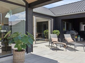 Ein modernes Haus mit einer Terrassenanlage, die zwei Lounge-Stühle, einen Grill und einen großen Kübel mit Pflanze zeigt.