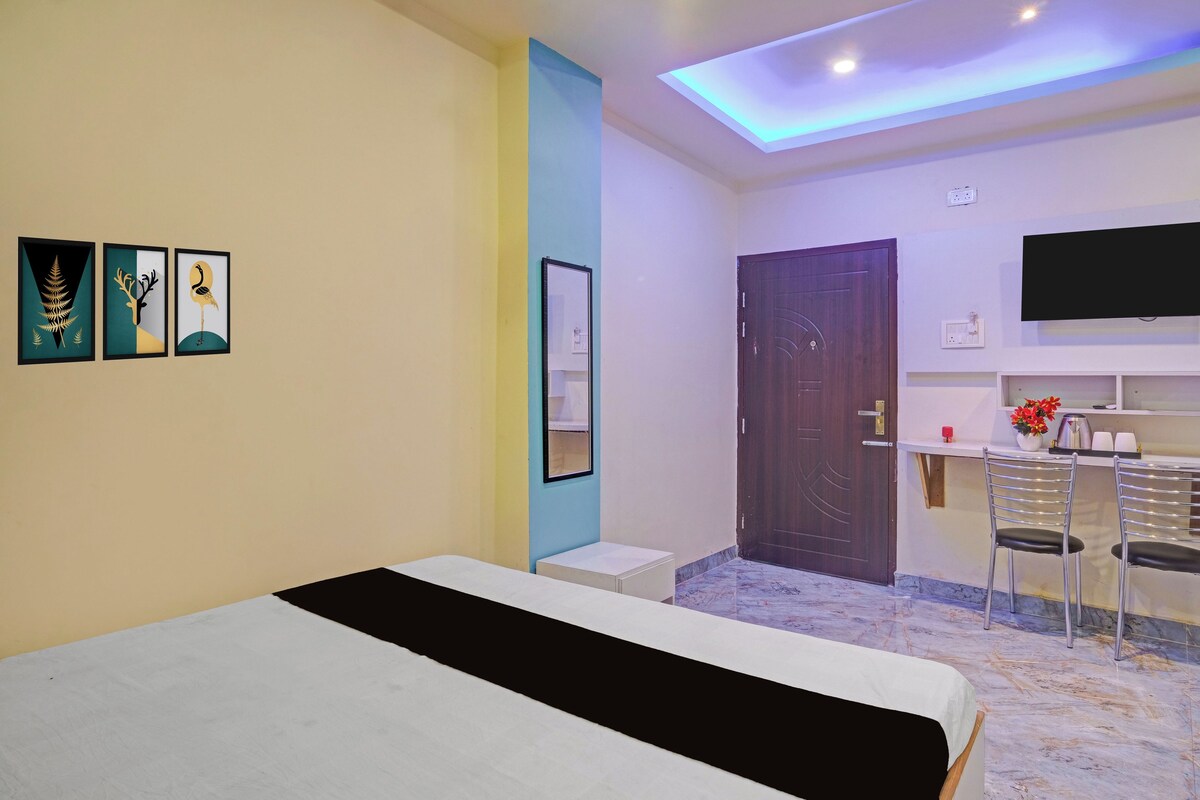 A bedroom with a bed, a desk with chairs, a TV, and artwork on the wall.