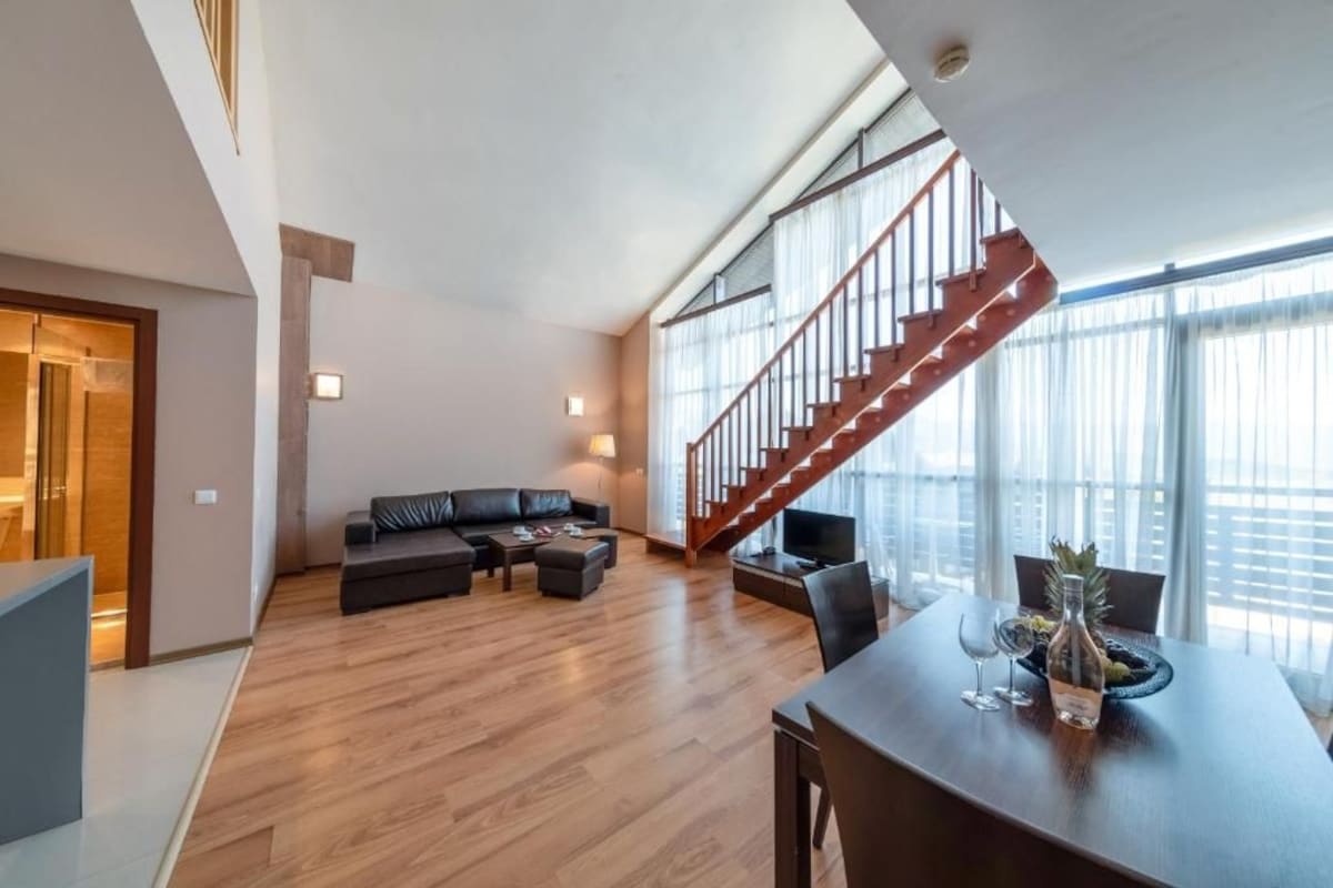 Una sala de estar moderna con piso de madera, un conjunto de sofá, una mesa de comedor con sillas y una escalera que lleva a un nivel superior.