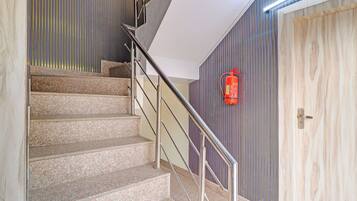 Una escalera con escalones de granito y barandillas metálicas, que conduce a una habitación con una puerta de madera y un extintor colgado en la pared.