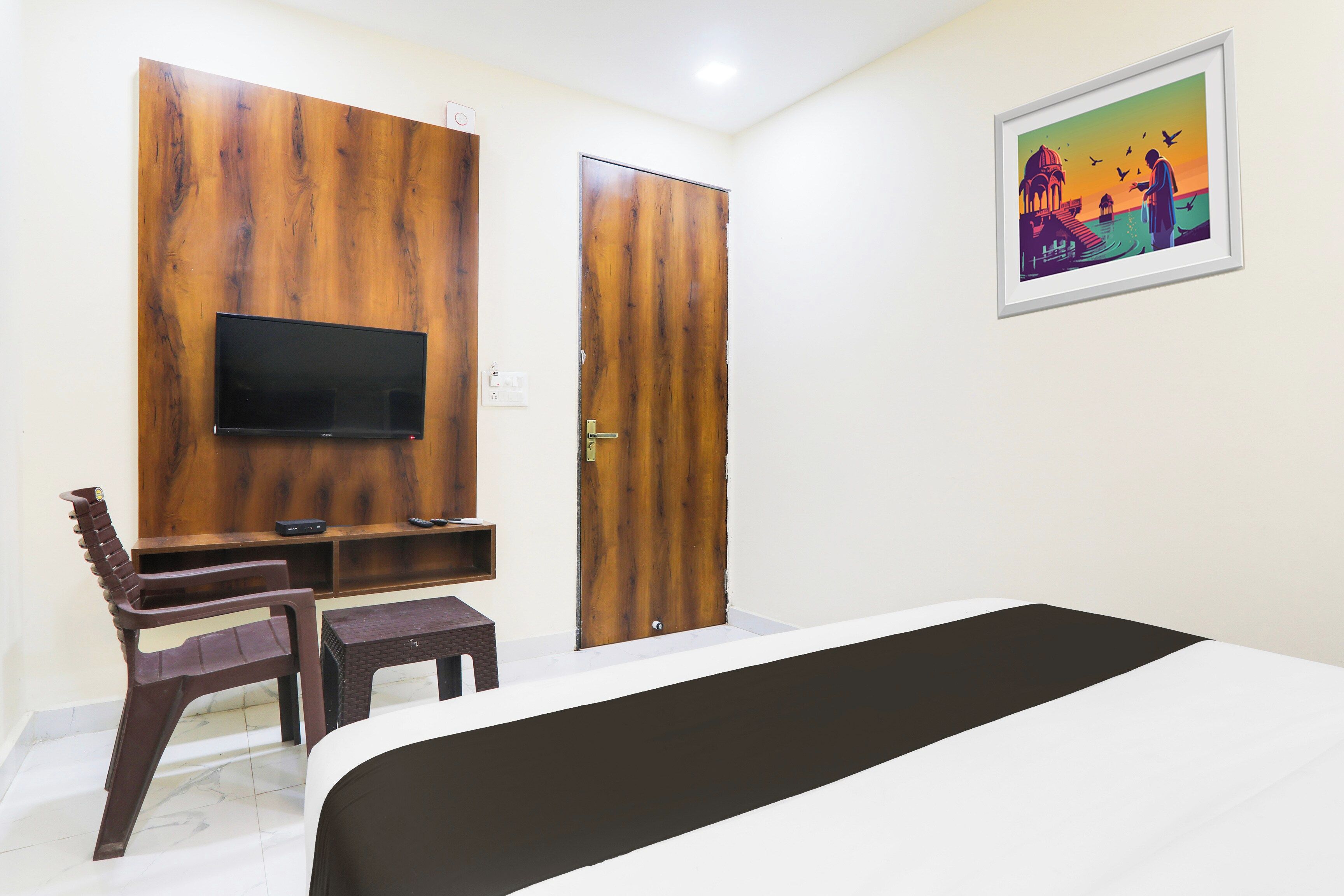 A hotel room with a bed, a TV mounted on a wooden panel, a desk with two chairs, and a door.