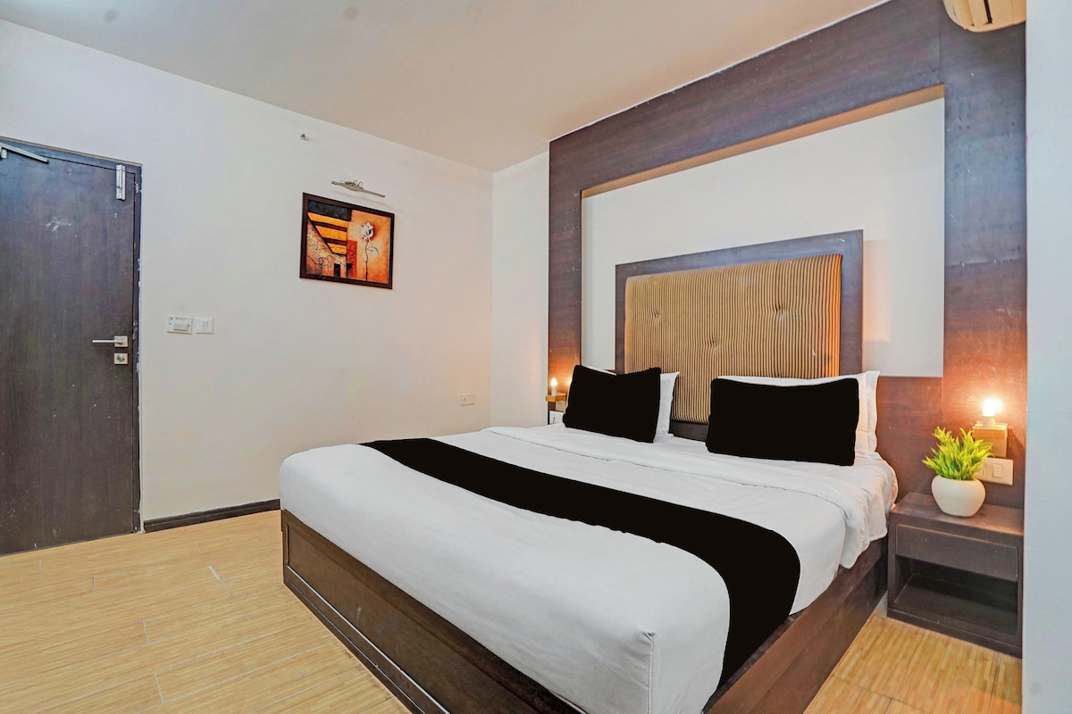 A hotel room with a large bed, a wooden headboard, a wall-mounted artwork, a bedside lamp, and a small potted plant.