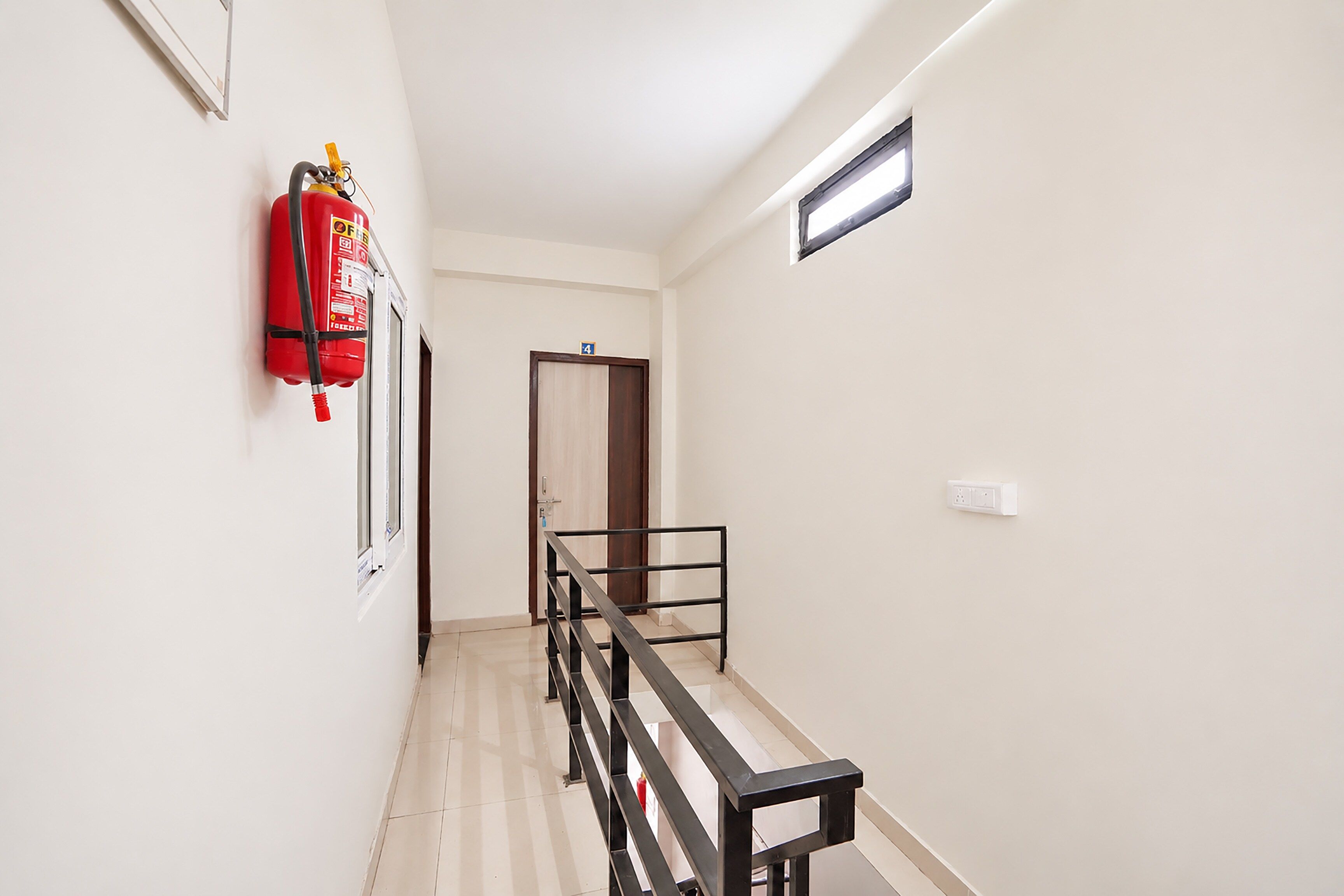 Un pasillo con un extintor en la pared, una puerta con un letrero de número y un ventanal con una rejilla.