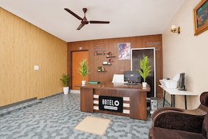 A hotel reception area with a wooden reception desk, a ceiling fan, a reception counter, a computer, a chair, and a potted plant.