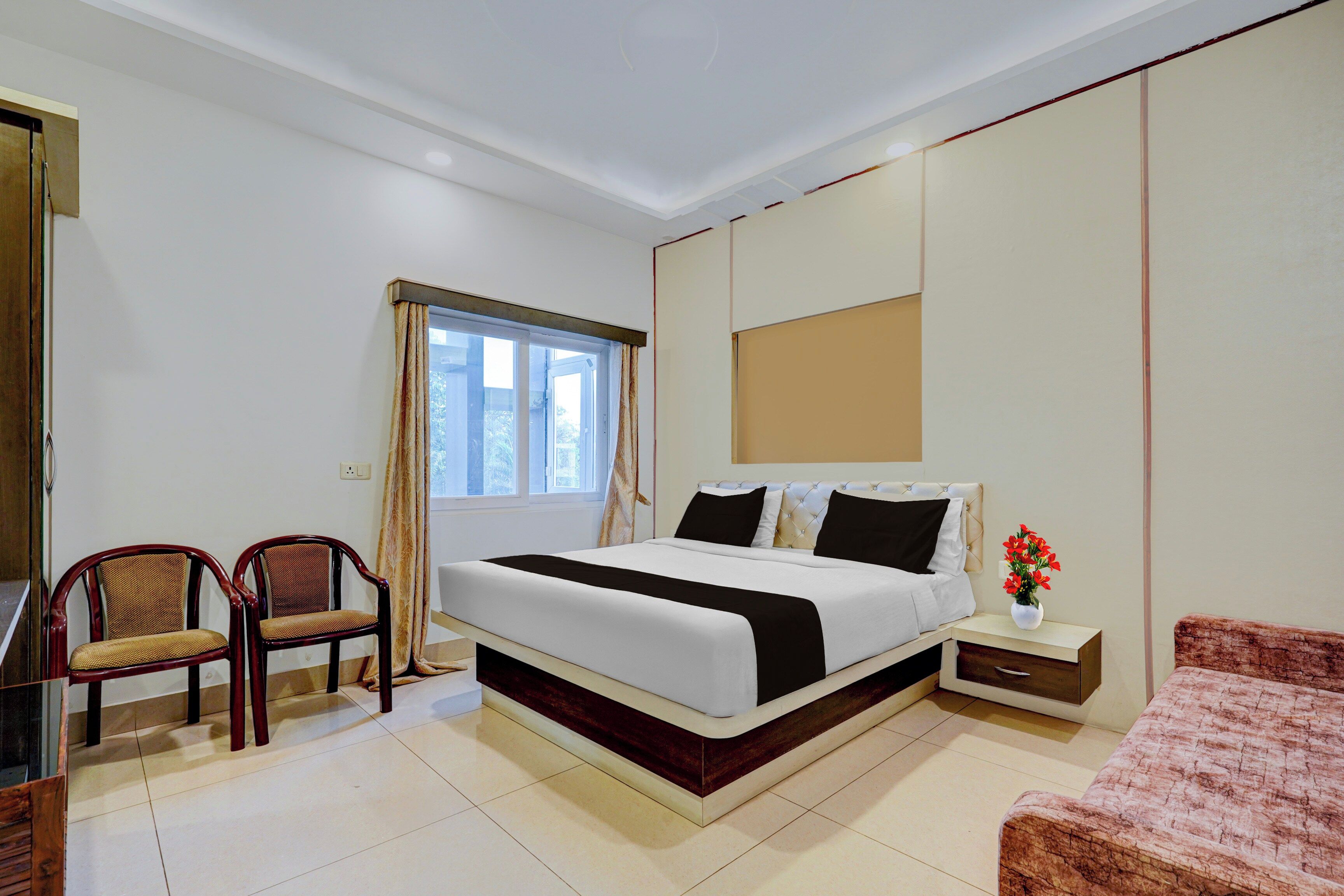 A hotel room with a bed, two chairs, a nightstand, a small table, and a window with curtains.