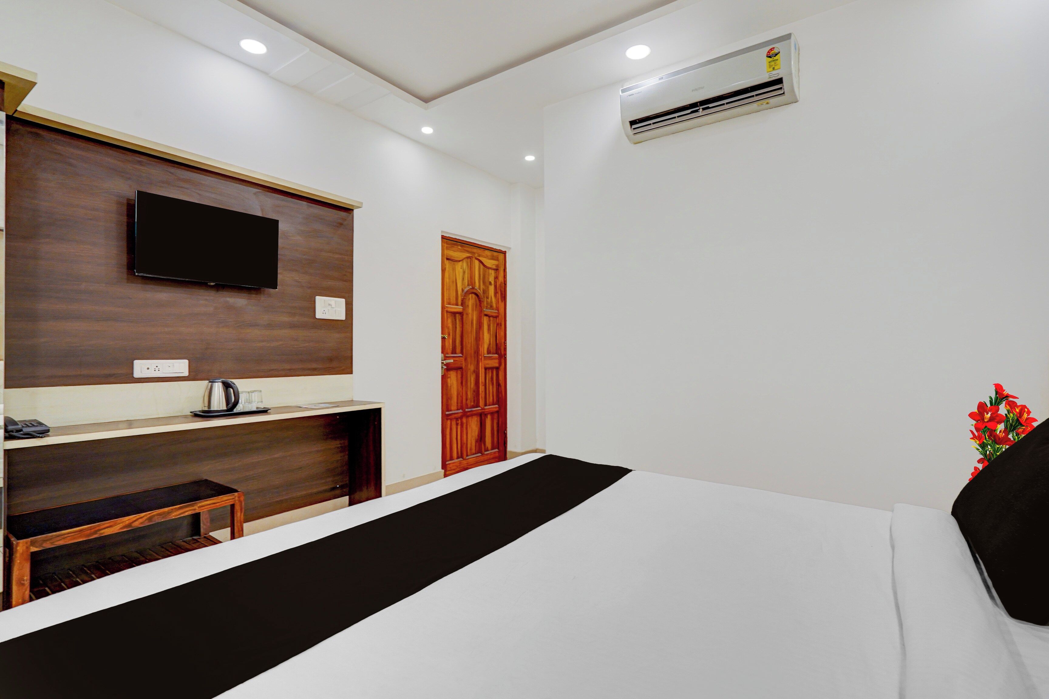A hotel room with a bed, a TV mounted on the wall, a wooden door, and an air conditioning unit.