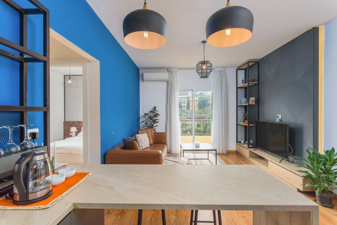A modern living room with a sofa, a coffee table, and a TV. There is a kitchen area with a kettle and a coffee machine. The room has blue walls and a large window.