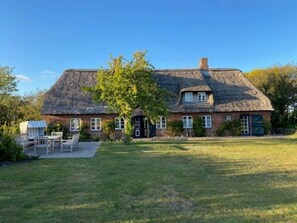 Ein reetgedecktes Haus mit Ziegelaußenwand, einer Haustür und einer kleinen Veranda mit Stühlen.