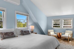 A bedroom with a large bed, two chairs, a lamp, and windows with a view of trees. - Cape Cod Escape: Sweeping Views & Outer Cape Adventures RPAM130 (Eastham)