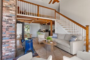 A spacious living area with a high ceiling, a staircase, and a kitchen in the background. - Cape Cod Escape: Sweeping Views & Outer Cape Adventures RPAM130 (Eastham)