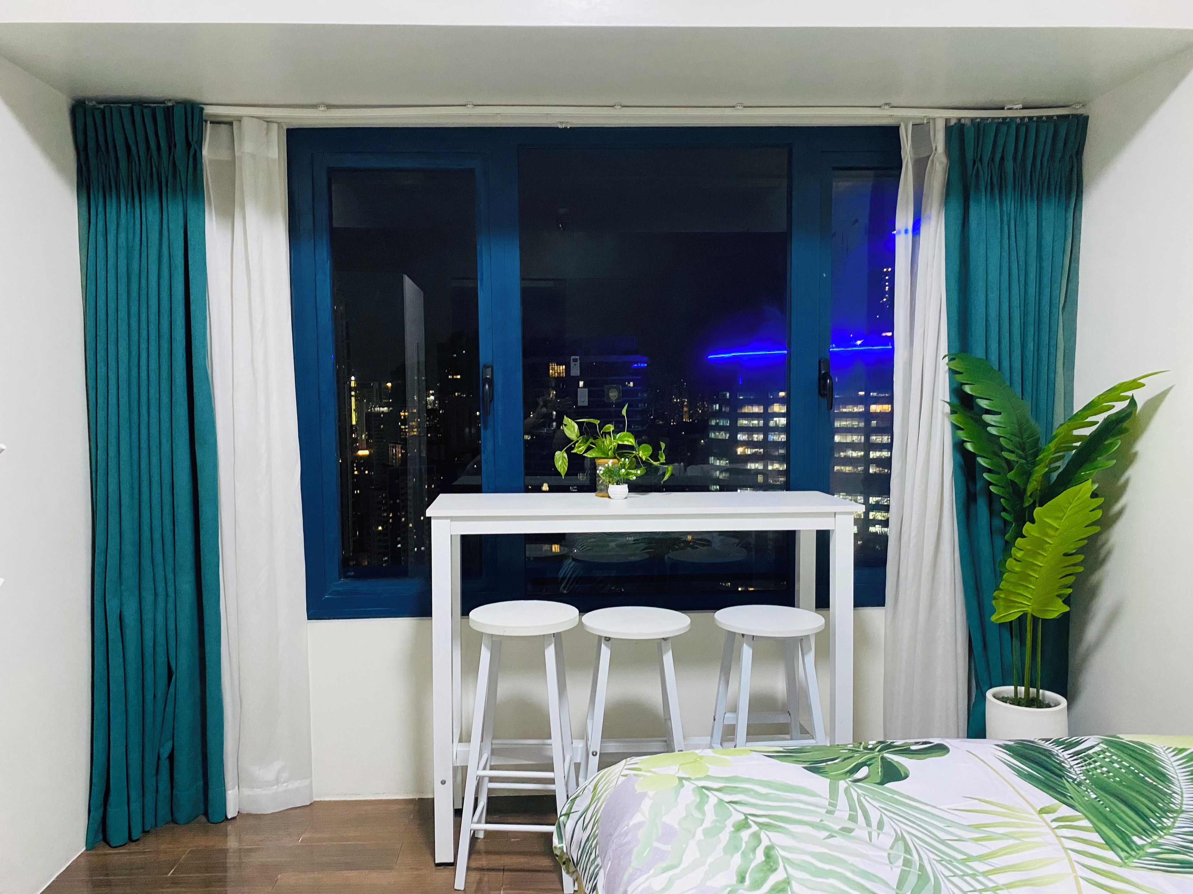 Una habitación con cama, una mesita con taburetes, una planta en maceta y un ventanal con cortinas.