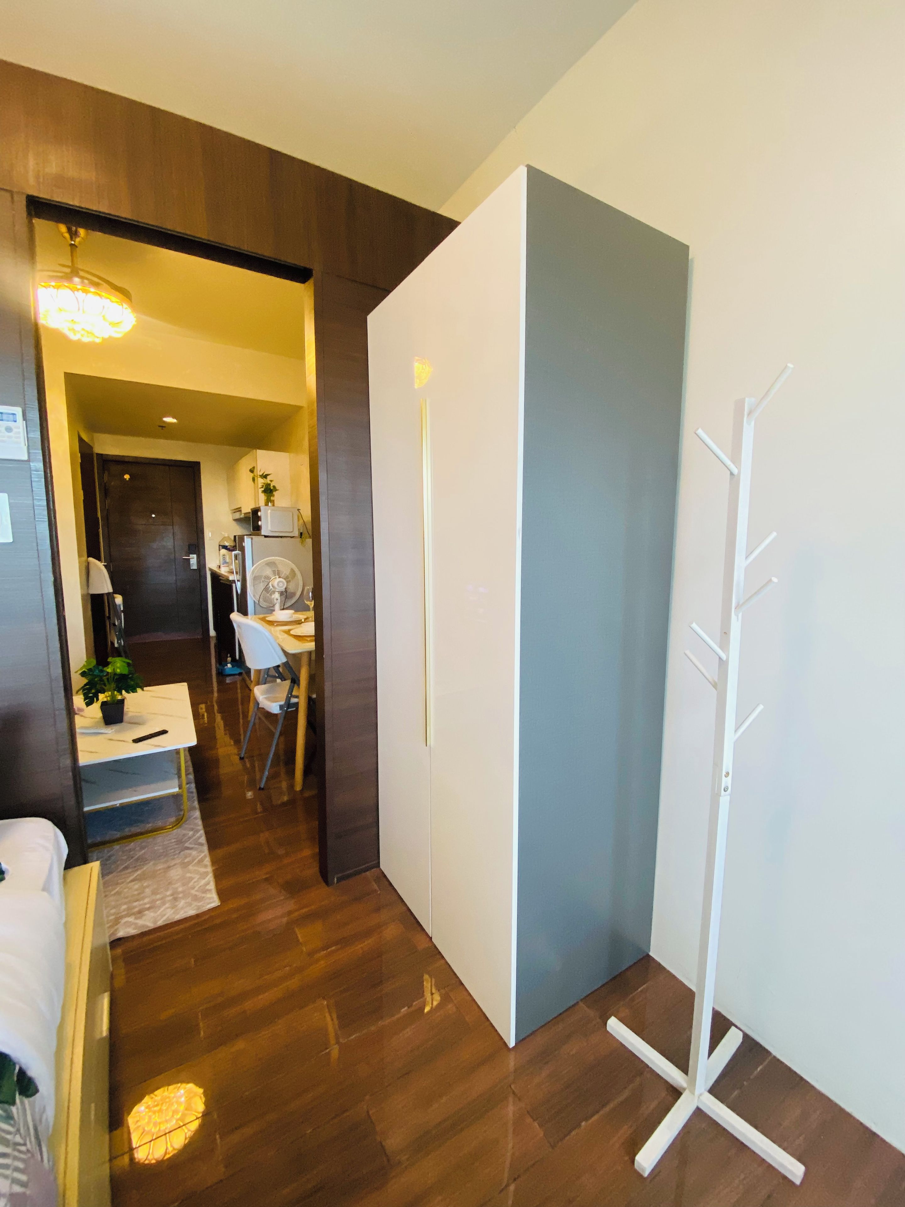 Habitación de hotel con piso de madera, cama blanca, escritorio con silla, perchero blanco y puerta que da a una zona de cocina.