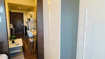 Habitación de hotel con piso de madera, cama blanca, escritorio con silla, perchero blanco y puerta que da a una zona de cocina.