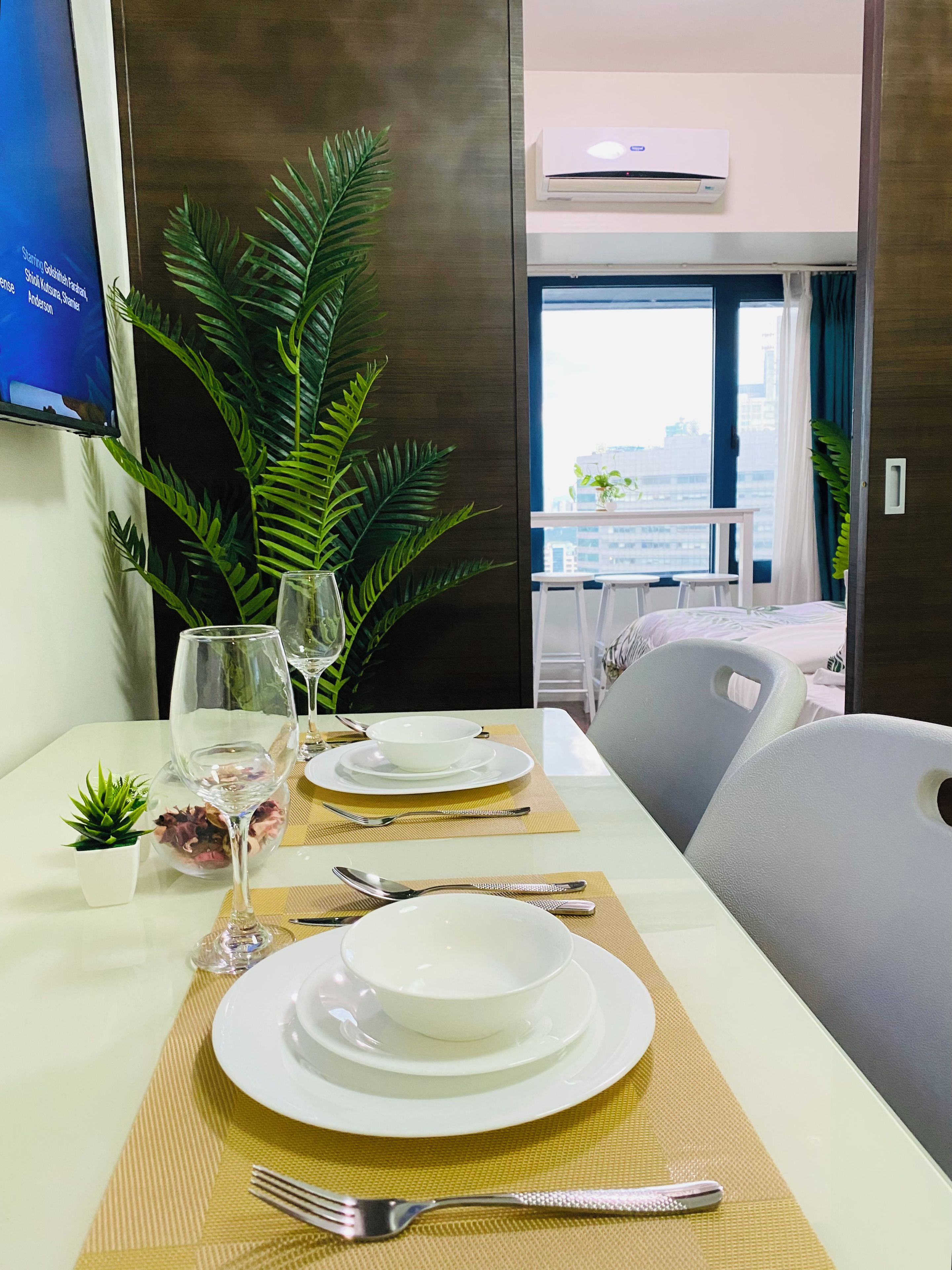 Habitación de hotel con una mesa de comedor para dos personas, una planta en maceta y vistas de la ciudad a través de la ventana.