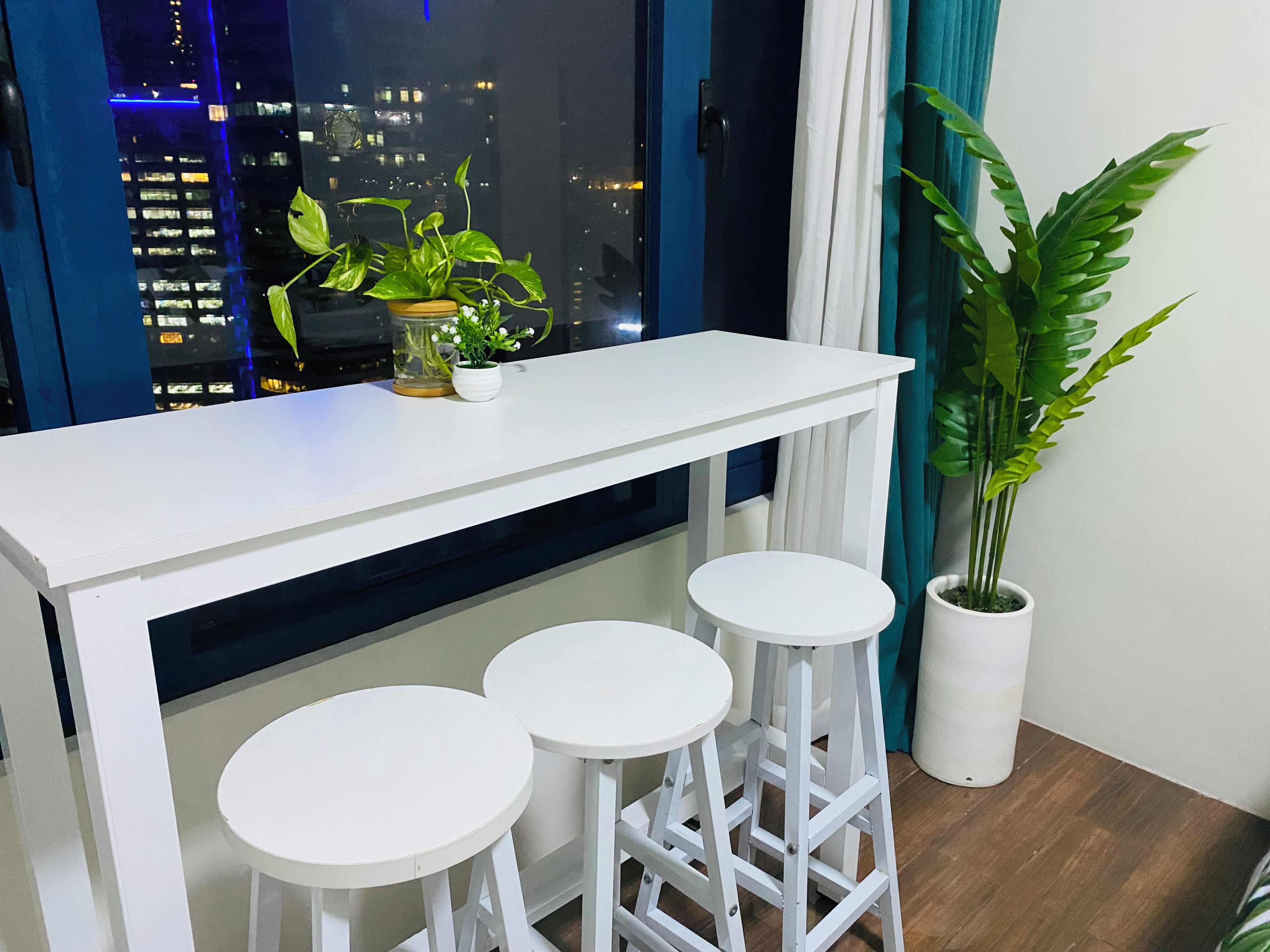 Un juego de taburetes blancos con una mesa blanca, una planta en maceta y una vista panorámica de la ciudad de noche.