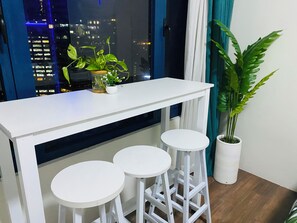 Un juego de taburetes blancos con una mesa blanca, una planta en maceta y una vista panorámica de la ciudad de noche.