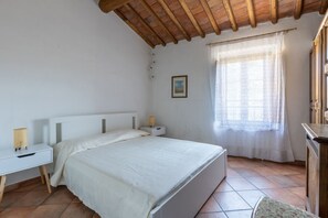 Un dormitori amb un llit, una tauleta de nit, una làmpada, una finestra amb cortina i un quadre a la paret.