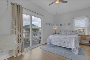A bedroom with a bed, a hanging chair, a ceiling fan, a window with blinds, and a view of a house outside. - The Bella Vista Beachfront Retreat + Pool, Elevator, Golf Cart & Ice Maker (Surf City)