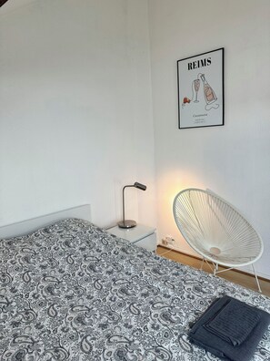 A bedroom with a patterned bedspread, a standing lamp, a chair, and a framed picture on the wall. - New Listing (Reims)