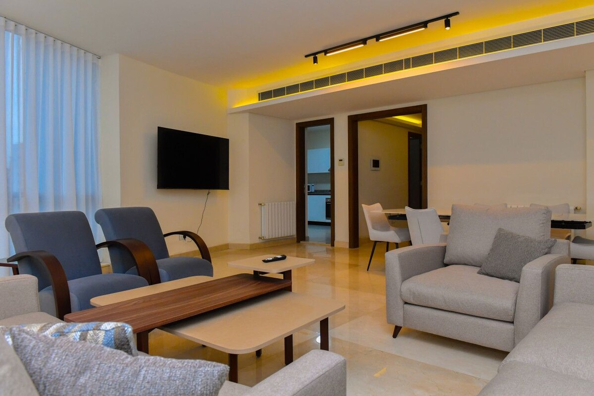A modern living room with a flat-screen TV, two sofas, a coffee table, and a dining area with chairs.