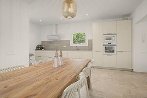 A modern kitchen with white cabinetry, a wooden dining table, and a pendant light. - Holiday Home 'Lo Tamariu' with Private Pool, Wi-Fi and Air Conditioning (Deltebre)