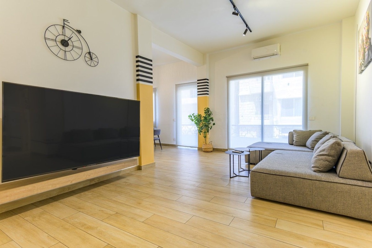 A modern living room with a flat-screen TV, a sofa, a coffee table, and a potted plant.
