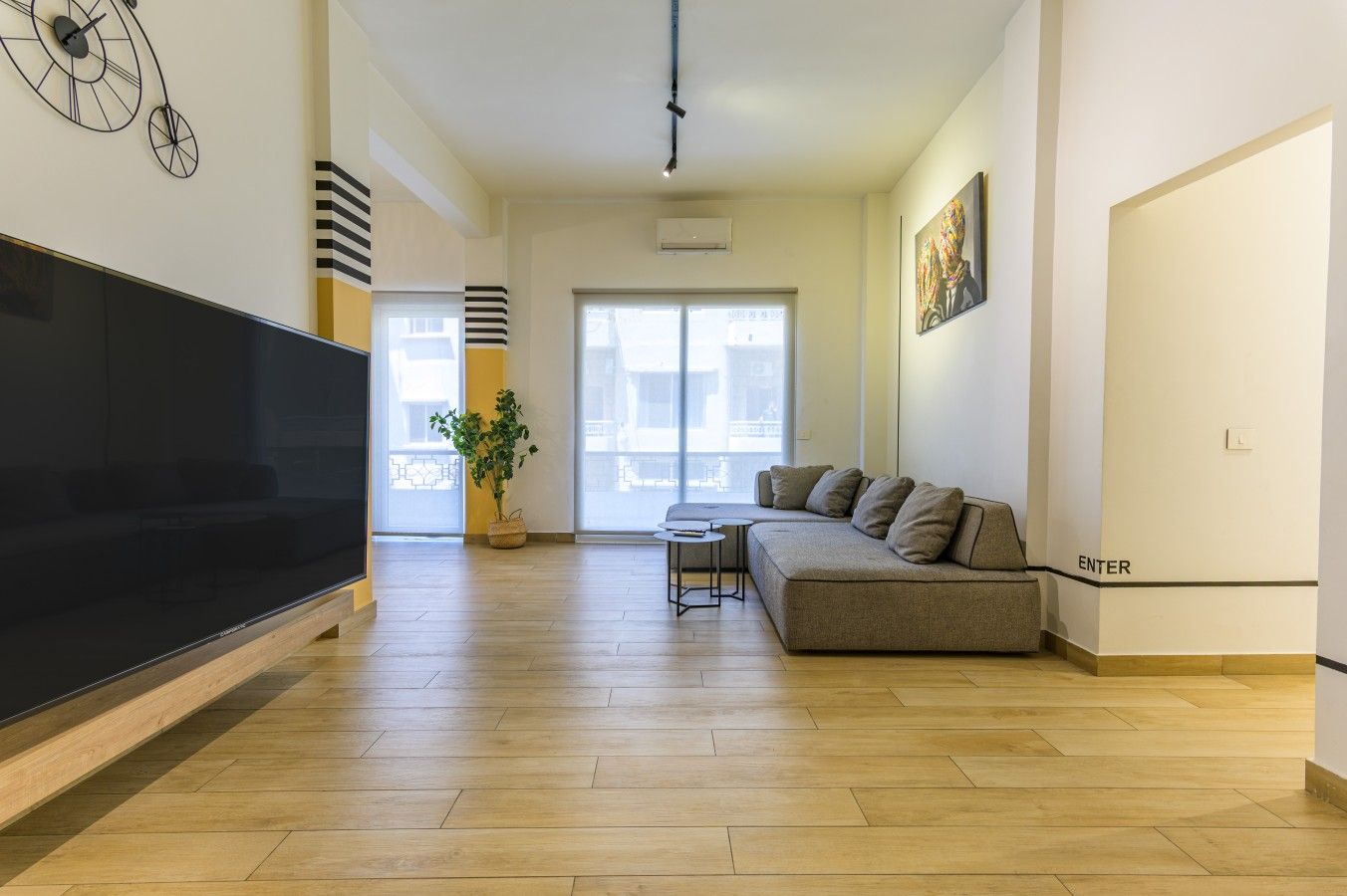 A modern living room with a large flat-screen TV, a grey sofa, a small coffee table, and a potted plant.