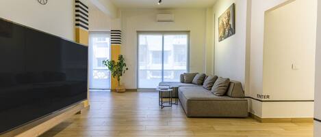 A modern living room with a large flat-screen TV, a grey sofa, a small coffee table, and a potted plant.