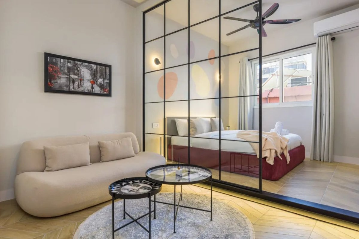 A modern hotel room with a sofa, a round coffee table, a bed with a red headboard, and a large window with curtains.