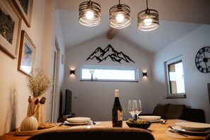 A dining area with a table set for two, a bottle of wine, and a bowl of grapes. There are three pendant lights above the table, a wall-mounted clock, and a framed picture on the wall.