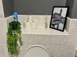A bathroom with a toilet, a shelf with toilet paper, a framed photo, and a hanging plant. - Oak & Noir 2 Bed 2 Bath Wifi Free Parking Near Uni (Manchester)