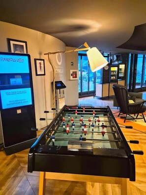Ein Kicker-Tisch in einem Raum mit einem digitalen Bildschirm, der Hotelinformationen zeigt, einer Lampe und Blick ins Freie.