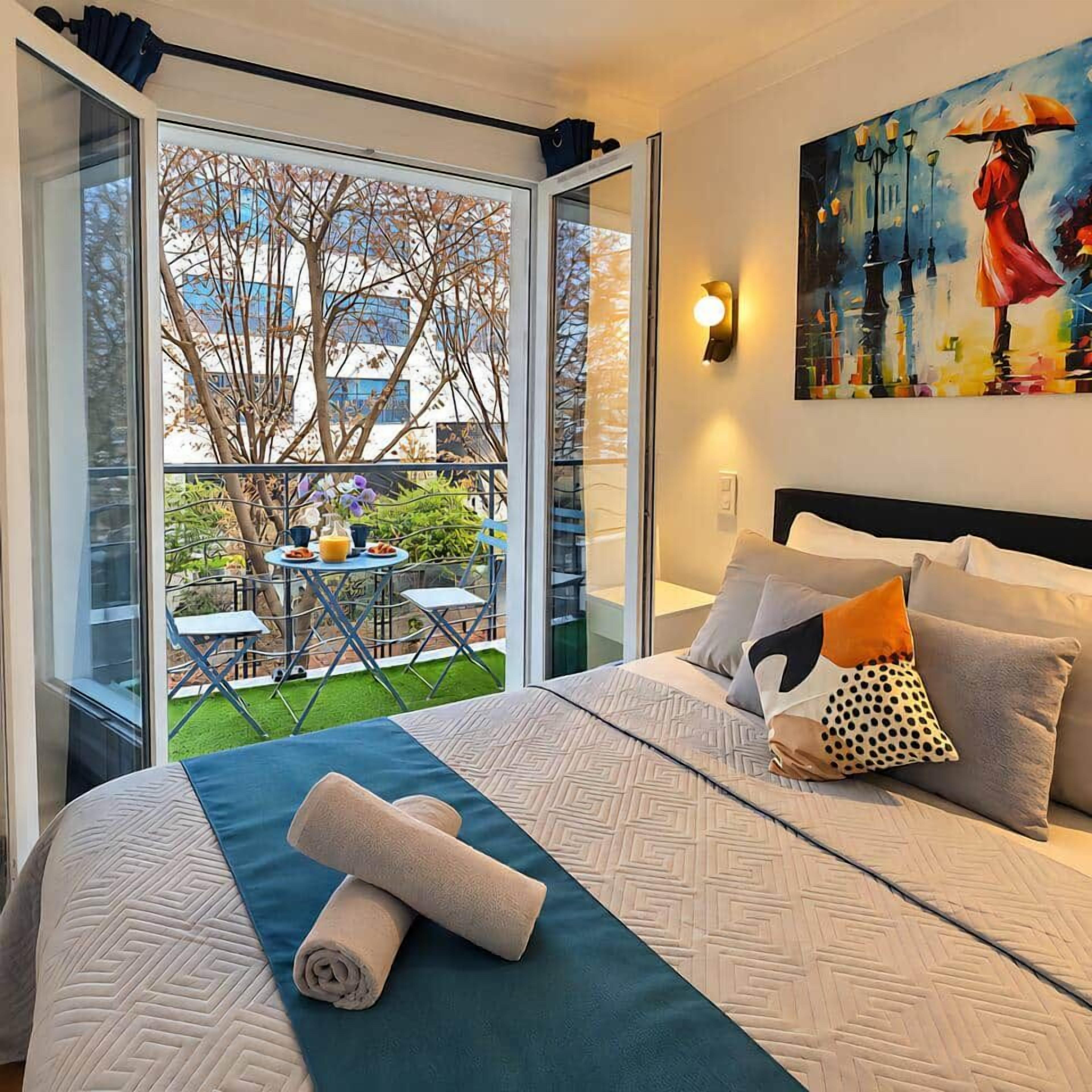 A hotel room with a bed, pillows, a rolled towel, a painting, and a view of the outdoors.