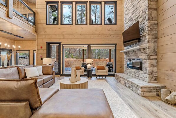 A spacious living area with a stone fireplace, leather sofas, and a large window overlooking a wooded area. - Heated Pool • Theater Room • Creekside Setting (Sevierville)