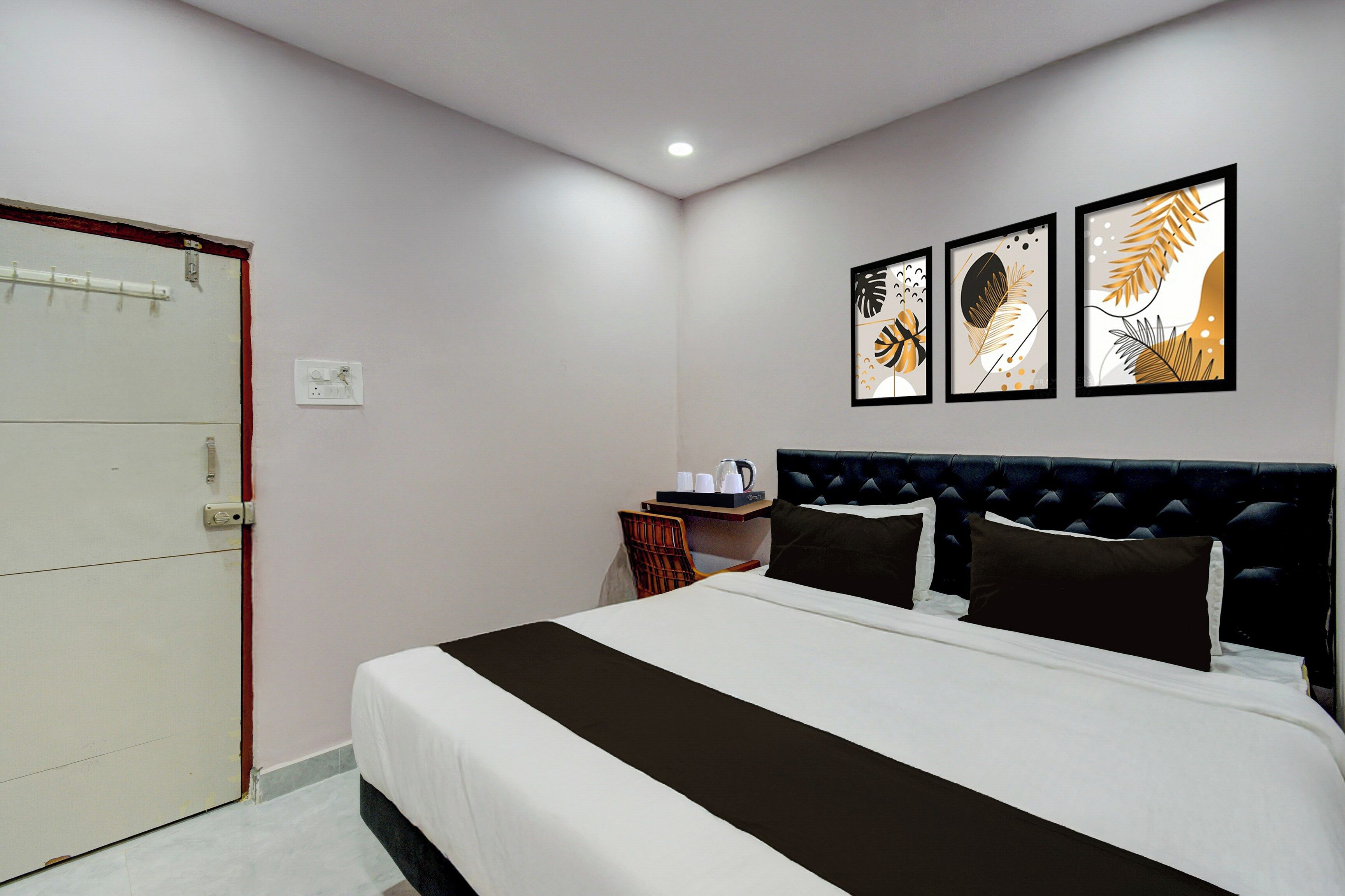 A hotel room with a bed, a bedside table with a kettle, and three framed artworks on the wall.