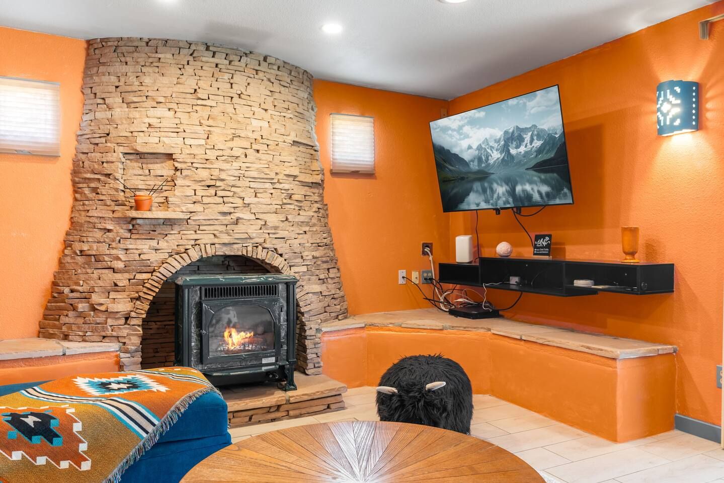 A room with a stone fireplace, a television mounted on the wall, and a black dog sitting on the floor.