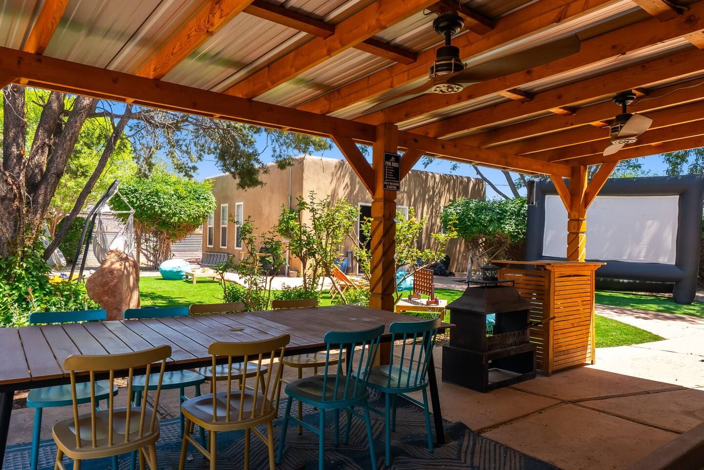 An outdoor patio with a wooden table and chairs, a barbecue grill, and a view of a backyard with a swing set and a house.