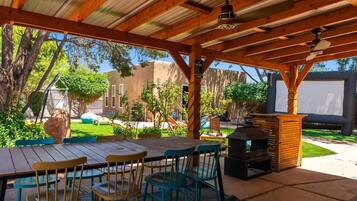 An outdoor patio with a wooden table and chairs, a barbecue grill, and a view of a backyard with a swing set and a house.