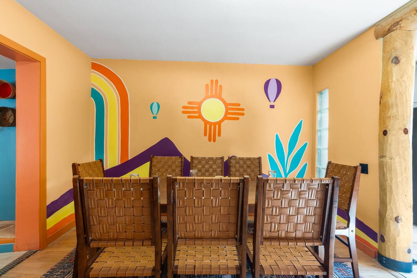 A room with a seating area featuring woven chairs, a wall with colorful murals, and a wooden pillar.