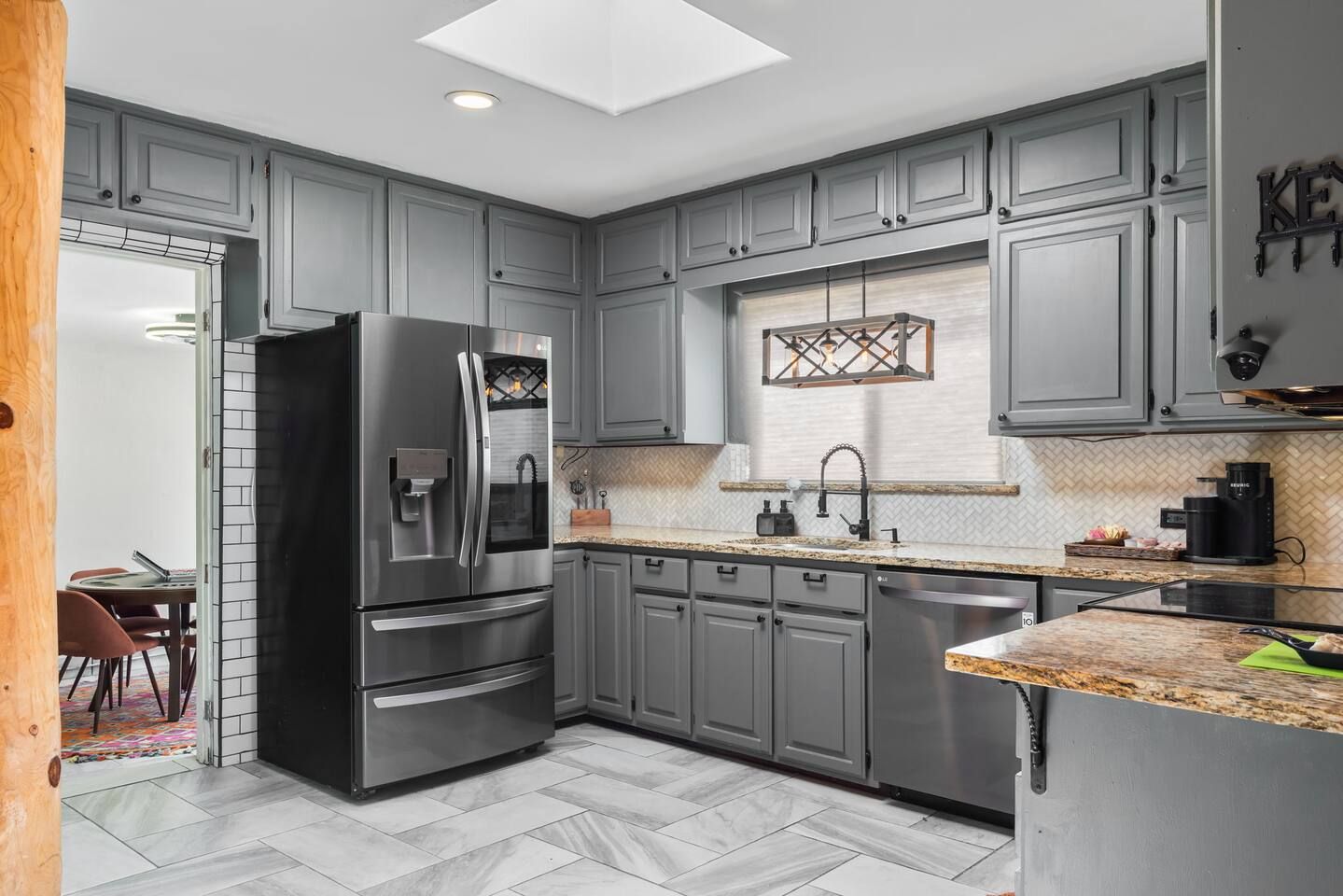 A modern kitchen with grey cabinets, a stainless steel refrigerator, a herringbone-patterned backsplash, and a skylight.