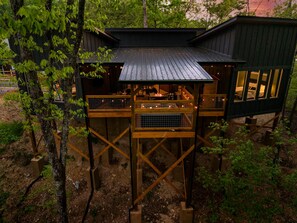 Ein Baumhaus mit einer Holzterrasse, einem offenen Bereich mit Feuerstelle und einem separaten, hüttenartigen Gebäude.