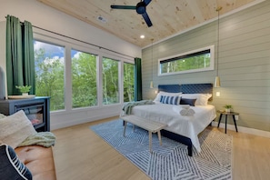 A bedroom with a large bed, a bench, a nightstand, and a view of trees through the windows. - The W Retreat • Sauna • Hot Tub • 2 King Suites (Sevierville)