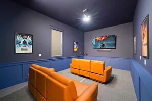 A home theater room with a blue wall, orange seating, a large TV, and movie posters. - Gulf Views-Pool-Beach 2 Min Walk-Hot Tub-Theatre (Santa Rosa Beach)