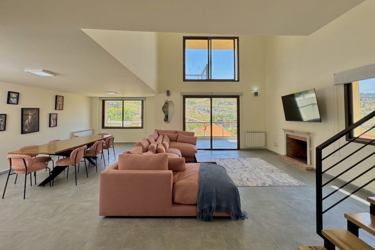 A spacious living room with a large sofa set, a dining table with chairs, a fireplace, and a TV mounted on the wall.