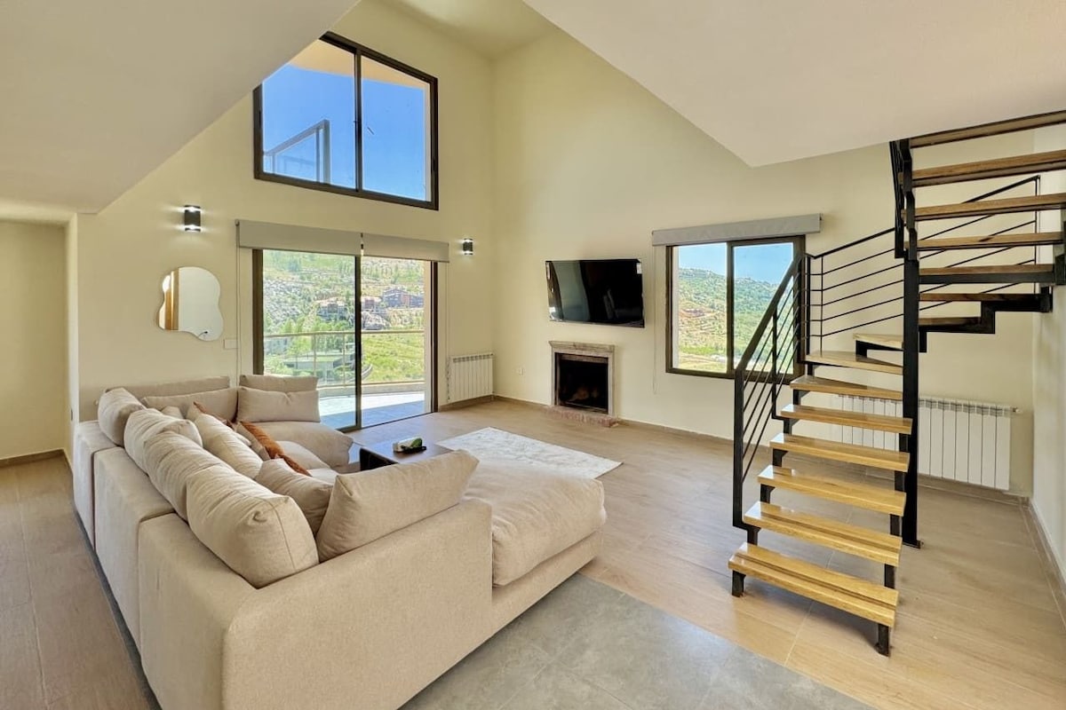 A spacious living room with a high ceiling, a large sofa, a fireplace, and a wooden staircase.