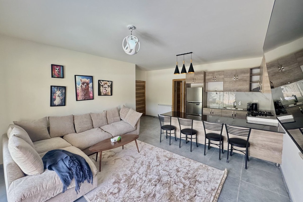 A modern living room with a sofa, coffee table, and a rug. There is a dining area with chairs and a kitchen with a stove and cabinets.