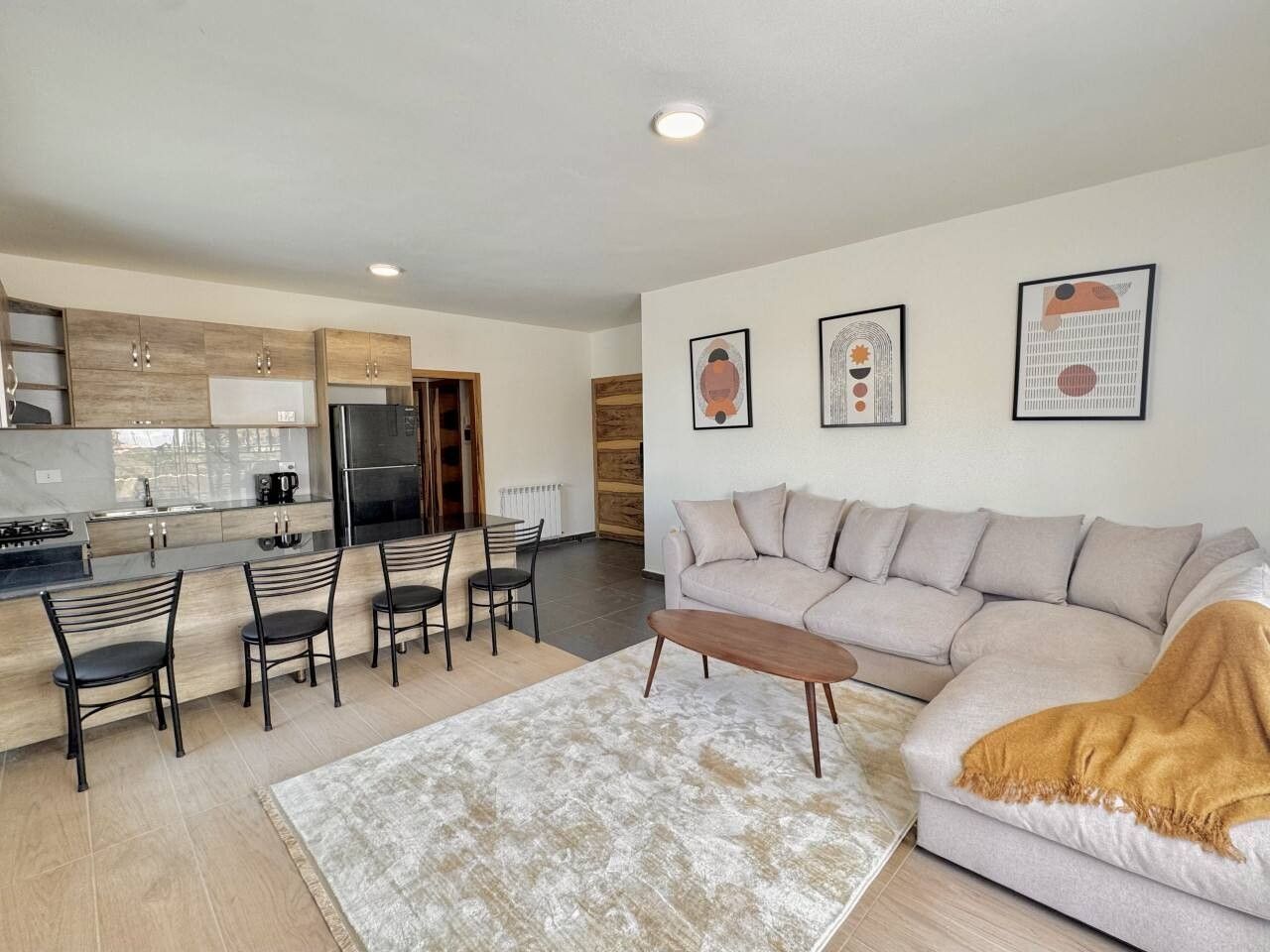 A modern living room with a sofa, coffee table, and chairs. There is a kitchen area with cabinets and appliances.