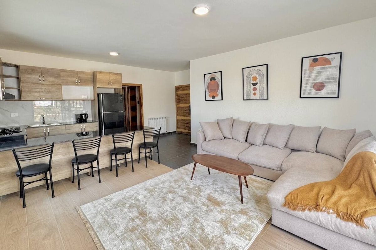 A modern living room with a sofa, coffee table, and chairs. There is a kitchen area with cabinets and appliances.